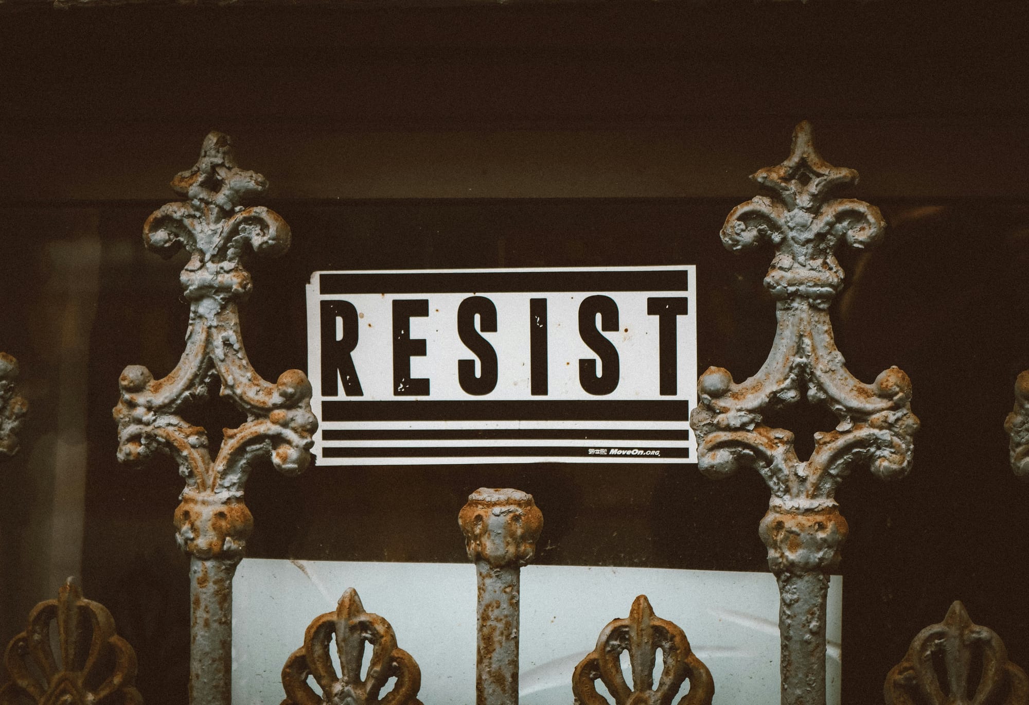 ornate, rusted iron fence in front of wall with black and white "RESIST" placard