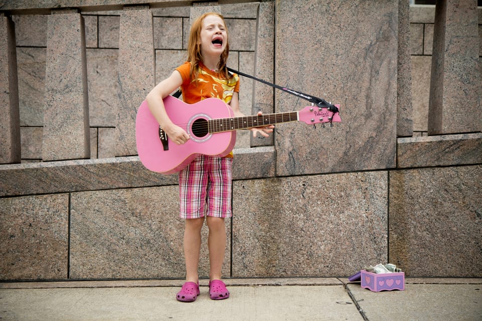 young red-haired girl standing on a street playing a pink guitar for tips