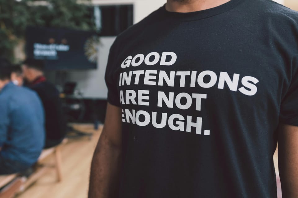 Black man from neck down wearing black t-shirt that says GOOD INTENTIONS ARE NOT ENOUGH.